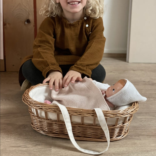 Mädchen sitzt vor dem Puppentragekorb aus Geflecht und deckt gerade die Puppe mit der weichen Decke zu.