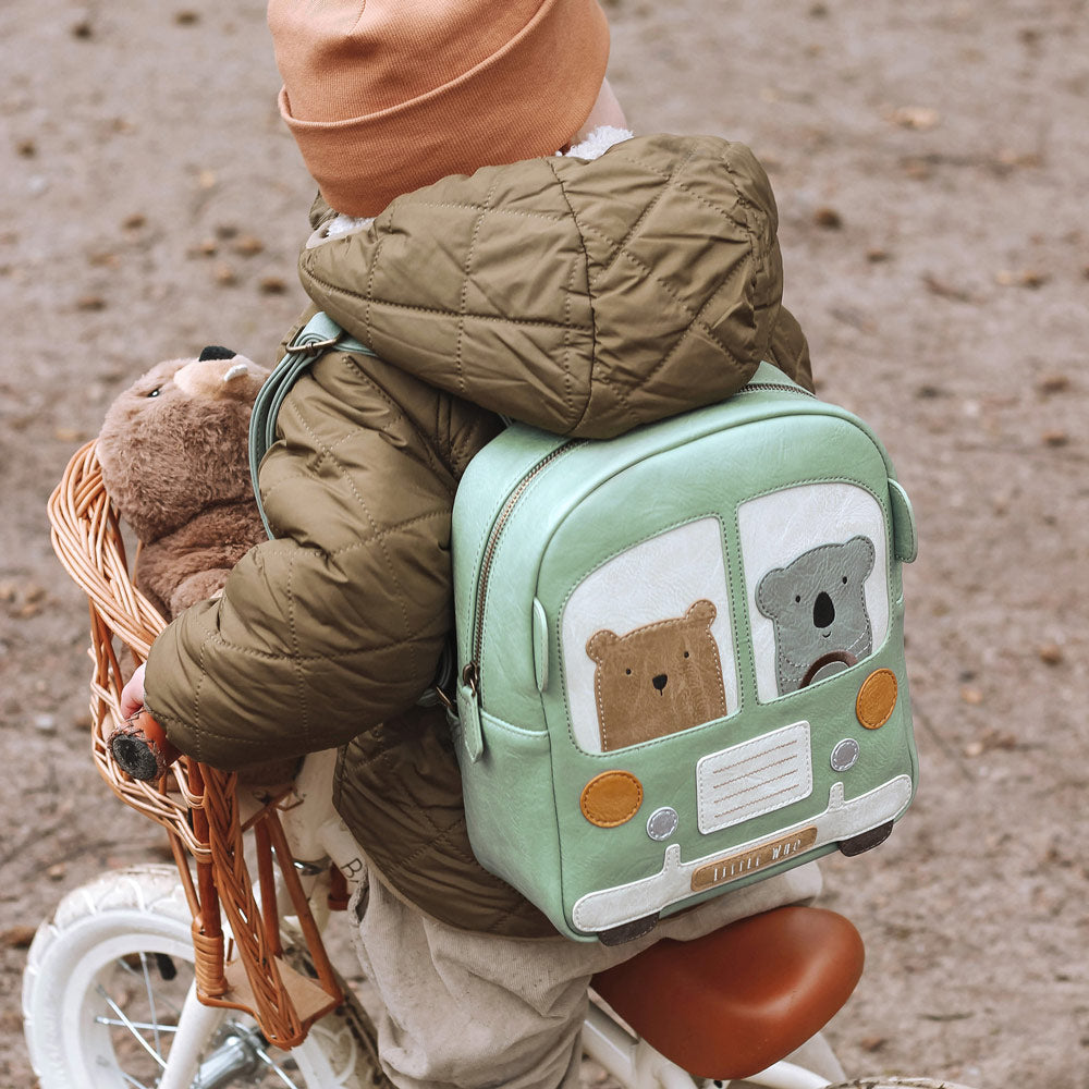 Kleinkind-Junge sitzt auf einem Fahrrad mit dem Rücken zur Kamera. Auf dem Rücken hat er den süßen Kinderrucksack an, der die Form eines Buses hat. Im Bus sitzen ein süßer Bär und ein Koala.