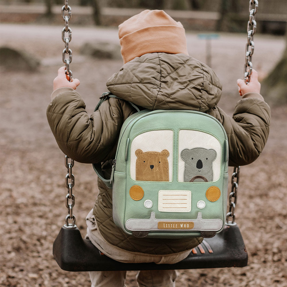 Kleinkind-Junge sitzt auf einer Schaukel mit dem Rücken zur Kamera. Auf dem Rücken hat er den süßen Kinderrucksack an, der die Form eines Buses hat. Im Bus sitzen ein süßer Bär und ein Koala.