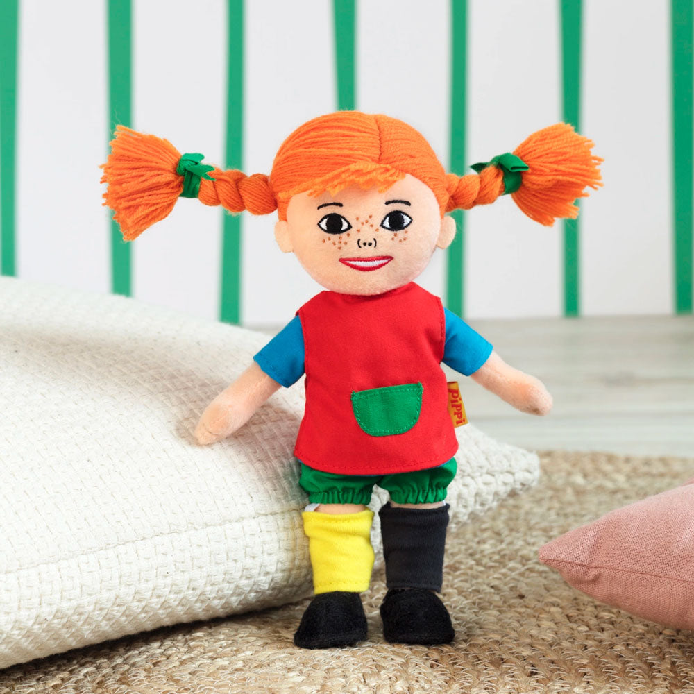 Pippi Langstrumpf Puppe hat ein aufgesticktes Gesicht mit Sommersprossen. Sie hat die typisch orangenen Haare zu zwei seitlichen Zöpfen mit grünen Schleifen im Haar. Sie trägt ein rotes Hemd mit blauen Ärmeln, grüner Bauchtasche, eine grüne Hose und zwei unterschiedliche Socken. Eine Socke ist gelb, die andere schwarz. Zudem trägt die Pippi Langstrumpg schwarze Schuhe.