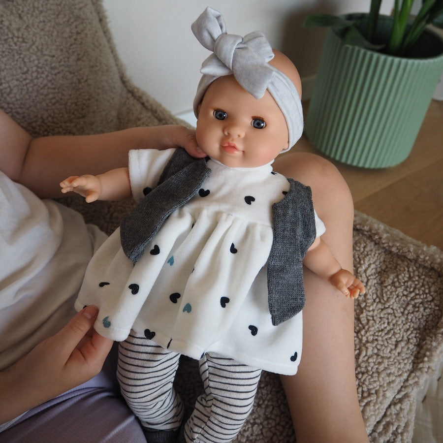 Die Puppe Agatha von Paola Reina trägt ein weißes Kleid mit Herzen drauf. Zudem trägt sie eine weiße Leggings mit grauen Streifen. Die Söckchen, das Stirnband und die Weste sind grau. Die Puppe hat blaue Augen, einen weichen Körper und Vinyl-Elemente.