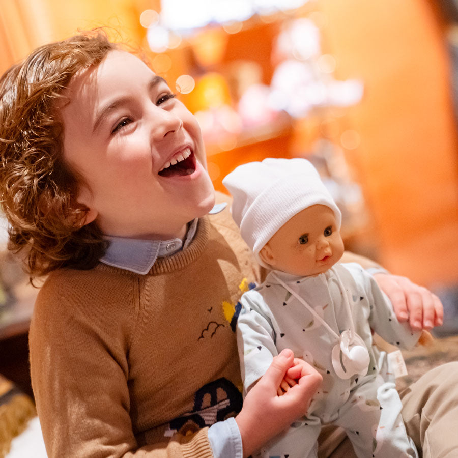 Junge freut sich mit der Babypuppe Martin, die den Strampler mit Möwen, einen Schnuller und eine weiße Mütze trägt.