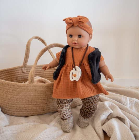 Kleidungsset für Puppe mit Leo-Hose, braunem Kleid und dunkler Weste. Puppe steht vor einem Puppentragebett. Den Schnuller trägt die Puppe um den Hals.