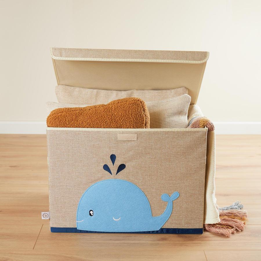 Die Aufbewahrungsbox ist rechteckig, ist in der Farbe Natur / beige und hat einen Wal aus Filz auf der Vorderseite. Der Wal hat die Farbe hellblau und spritzt dunkelblaue Tropfen. In der Box liegen Kissen und Decken.