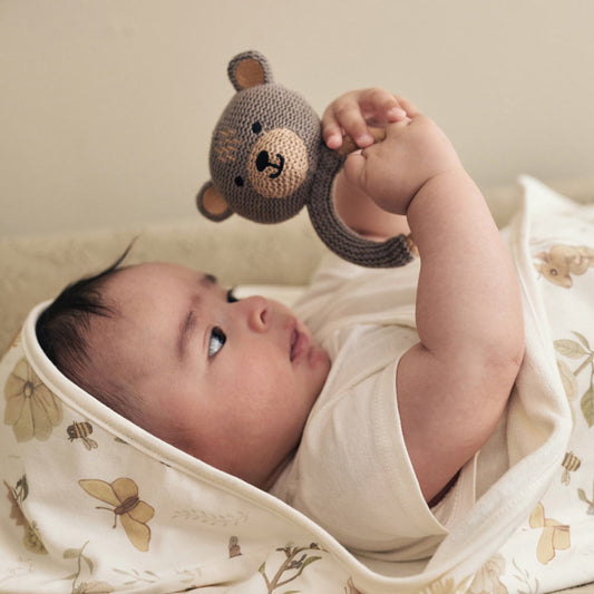 Baby hält Beißring mit den Händen und schaut auf den braunen Teddybär 