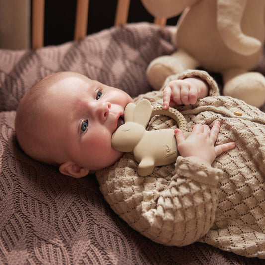 Baby liegt im Babybett und beißt auf dem Beißring Hase rum. Der beißring ist in der Farbe Nougat und sieht richtig süß aus. 