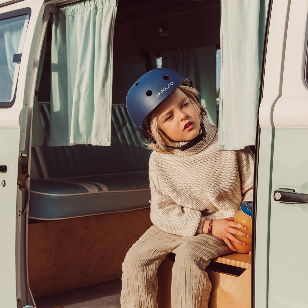 Junge mit blonden Haare sitzt in einem Camper und macht eine Trinkpause. Seinen naviblauen Helm von Banwood trägt er noch auf dem Kopf.