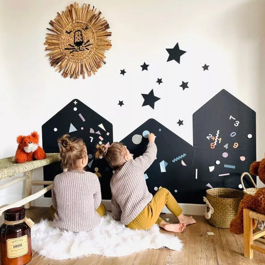 Zwei Kleinkinder spielen an den Magnettafel-Häusern mit Magneten. Die drei Häuser sind ebeneinander an der Wand geklebt. Die Sterne kleben dekorativ über den Häusern.