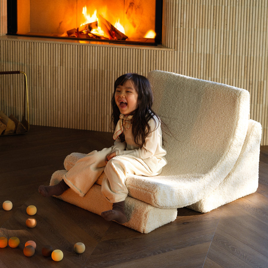 Kind sitzt im Wohnzimmer vor dem Kamin auf dem Moon Chair in der Farbe creme und lacht. 