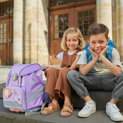 Zwei Kinder sitzen vor der Schule, der Junge trägt den Schulranzen in blau, das Mädchen sitzt neben ihrem Schulranzen in der Farbe lila. 