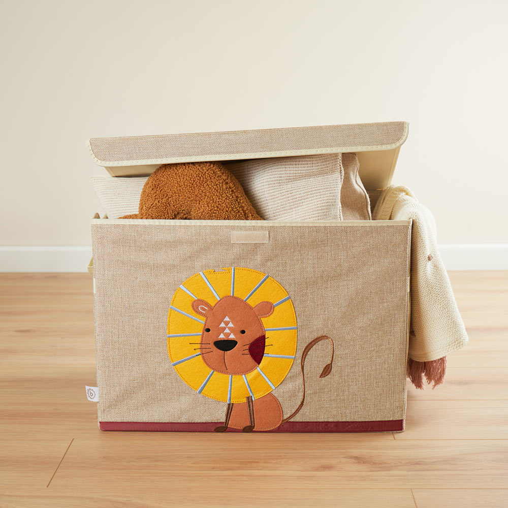 Die Aufbewahrungsbox ist rechteckig, ist in der Farbe Natur / beige und hat einen Löwen aus Filz auf der Vorderseite. Der Löwe hat eine gelbe Mähne und einen orangenen Körper. In der Box liegen Decken und ein Kissen.