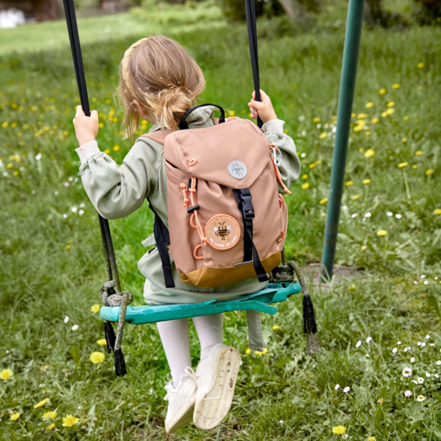 Mädchen auf der Schaukel trägt den Wanderrucksack in der Farbe braun. 